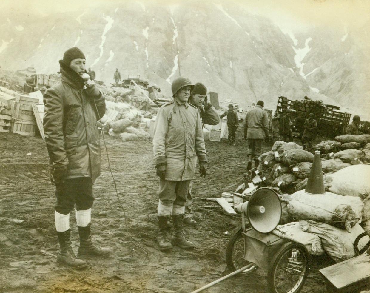 Loudspeaker Guides Landing Troops. ATTU - Landing boats bringing American troops to the rocky shores of Attu, westernmost island of the Aleutian chain, receive their instructions...through a loadspeaker. Standing on shore amid...piles of crates and bags filled with supplies...and ammunition, this Navy officer broadcasts...commands to landing troops. Latest reports...Attu indicate that the island's rough...may delay the final American push...;