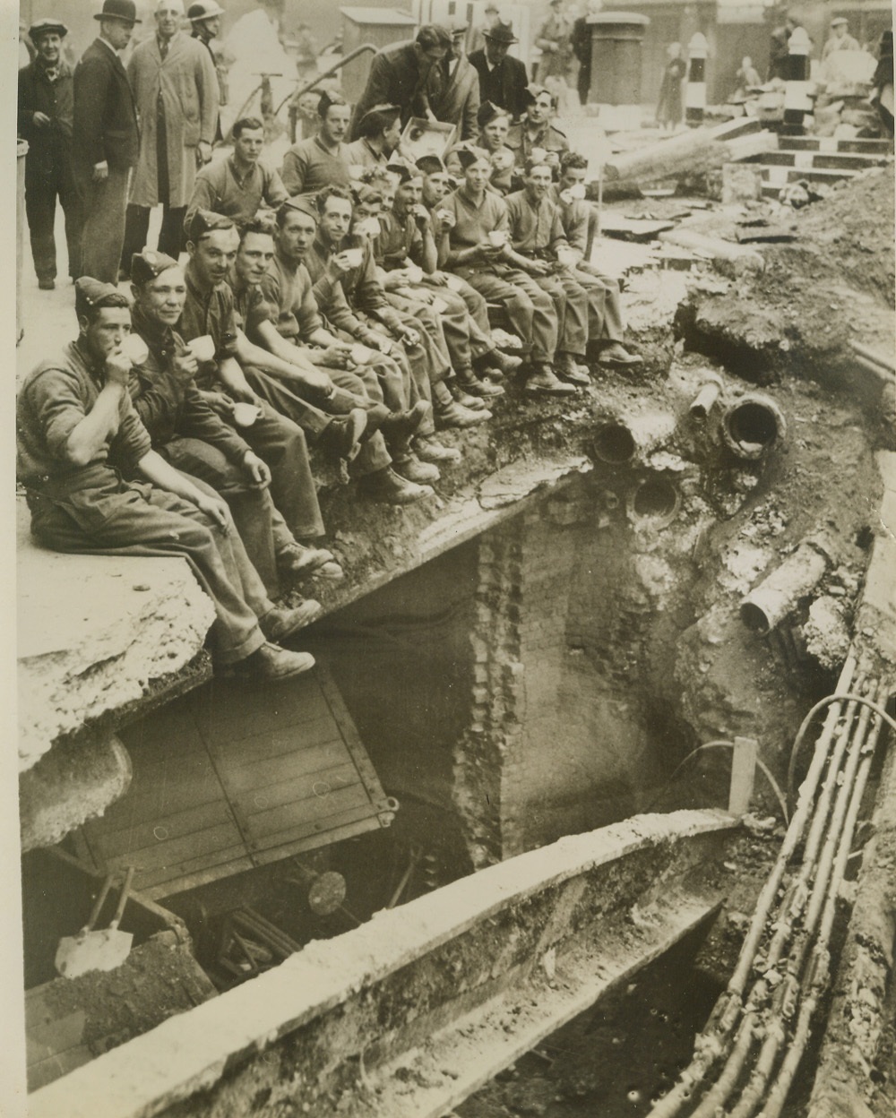 Army Pioneers Rest From Labors in London, 10/21/1940  LONDON, ENGLAND -- Sitting on the edge of a bomb crater these men of the Auxiliary Military Pioneer Corps refresh themselves with buns and tea before tackling their great task of clearing away wreckage caused by German raiders. Evidently this bomb blew the top off an underground railway. The end of a freight car and twisted rails can be seen in the left foreground. The Government has ordered 5,000 of these men into London to help the work of clearing away wreckage. Credit: (ACME);